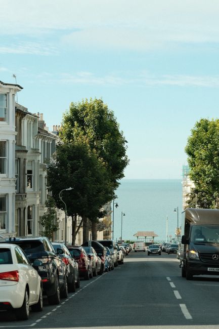A residential street scene during daytime showing a row of parked cars on both sides of the road, including various models and colours, with a large tree in the foreground partially obstructing the vi