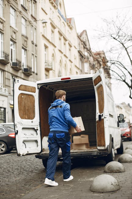 A man wearing glasses and a light blue shirt is seated on a wooden chair next to a white moving van parked outside a modern building with large glass windows. The van’s rear doors are open, revealin