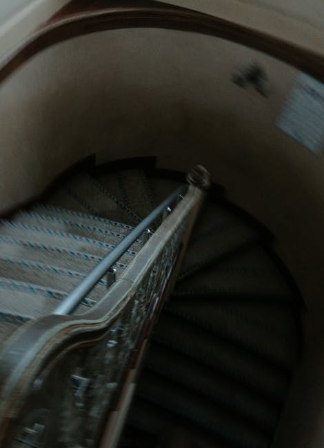 Inside a narrow staircase with wooden banisters and a curved wall, a team from Man with Van St Helier is engaged in a home relocation process. Several cardboard boxes, wrapped in plastic and secured with packing tape, are positioned on the staircase steps and along the landing area, indicating packing and moving preparations. A metal handrail runs alongside the staircase, assisting safe navigation during the loading process. The steps are carpeted, and natural light filters through a nearby window, illuminating the scene. Wooden furniture and large household items are either being carried or prepared for transport, with some objects close to the staircase entrance. The environment suggests a careful, organized furniture transport operation typical of professional removals, focusing on facilitating a smooth and secure move within the property for a residential customer, as part of comprehensive packing and loading services offered by Man with Van St Helier.