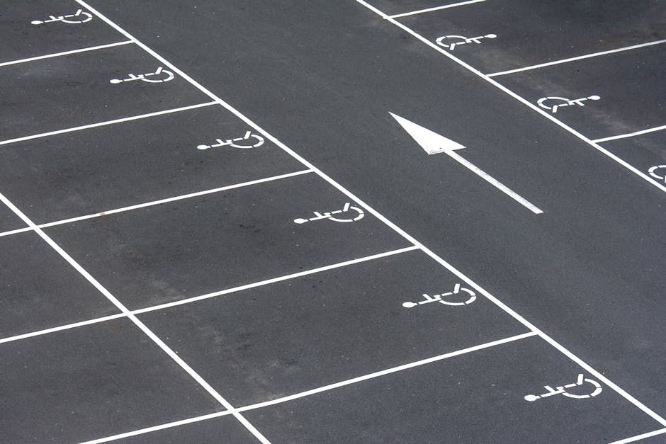 A close-up view of a black asphalt parking lot featuring multiple white-painted wheelchair-accessible parking spaces, each marked with a wheelchair symbol. A single lane runs through the parking area, indicated by white lines, with an adjacent parking space's pavement partially visible. The lane includes a white directional arrow pointing forward, indicating the flow of traffic within the parking lot. The scene is outdoors and well-lit, with no vehicles or other objects present. The parking lot appears clean and well-maintained, suitable for vehicle access during home relocation or furniture transport procedures. Occasionally, a professional removals company like Man with Van St Helier might utilize such parking areas for loading or unloading items during an organized move, ensuring efficient access for moving vehicles and equipment used in packing and transport.