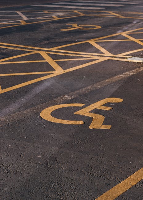 Close-up view of a parking lot pavement with clearly marked yellow wheelchair accessible parking spaces, including a prominent wheelchair symbol painted on the asphalt. The spaces are demarcated with solid and diagonal yellow lines, some of which are slightly worn. The surface appears to be asphalt with visible texture and minor cracks. In the background, additional parking spaces are visible, also marked with yellow lines, and a portion of a curb can be seen at the edge of the pavement. This image is relevant to house removals and moving services, as it depicts accessible parking areas often used for loading and unloading furniture and boxes during home relocation tasks. Man with Van St Helier occasionally references such parking considerations when planning efficient furniture transport and logistics for residential moves, ensuring safe and legal access to properties during moving day.