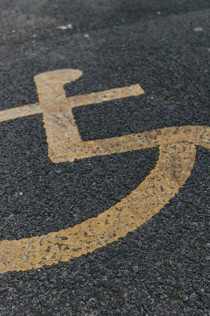 A close-up view of a black asphalt parking lot featuring multiple white-painted wheelchair-accessible parking spaces, each marked with a wheelchair symbol. A single lane runs through the parking area, indicated by white lines, with an adjacent parking space's pavement partially visible. The lane includes a white directional arrow pointing forward, indicating the flow of traffic within the parking lot. The scene is outdoors and well-lit, with no vehicles or other objects present. The parking lot appears clean and well-maintained, suitable for vehicle access during home relocation or furniture transport procedures. Occasionally, a professional removals company like Man with Van St Helier might utilize such parking areas for loading or unloading items during an organized move, ensuring efficient access for moving vehicles and equipment used in packing and transport.