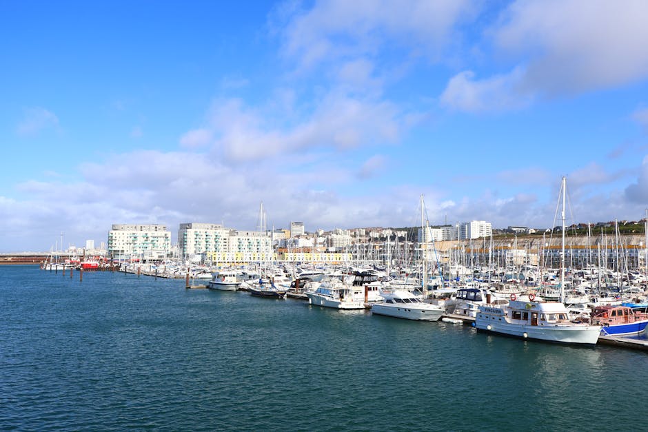 A marina with multiple white and blue motorboats and sailboats moored along wooden docks, with their masts rising vertically and some covered with protective fabric. Behind the boats, there are modern white residential buildings with large windows, set against a partly cloudy sky with blue patches. The water in the marina is calm and reflects the vessels and buildings, indicating a peaceful waterside environment. This scene depicts a typical location where house removals and furniture transport services by Man with Van St Helier might prepare for or conduct a home relocation, leveraging loading areas and access to water transportation for efficient moving processes.