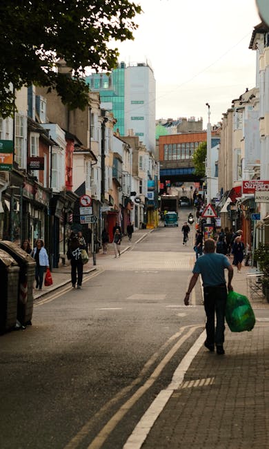 A street scene in St Helier showing a man walking away from the camera carrying a large green bag in his right hand, while facing a busy urban area lined with shops and cafes on both sides. The street features a mix of older and modern buildings, some with shop fronts and signage, and a few pedestrians walking along the sidewalks. In the background, taller buildings and a partly cloudy sky are visible. This scene illustrates a typical day in the town center, relevant to home relocation and furniture transport processes handled by Man with Van St Helier, situated within the context of household removals and moving services.