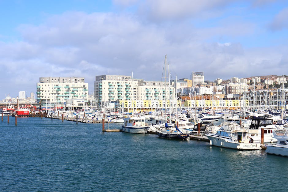 A street scene in St Helier showing a man walking away from the camera carrying a large green bag in his right hand, while facing a busy urban area lined with shops and cafes on both sides. The street features a mix of older and modern buildings, some with shop fronts and signage, and a few pedestrians walking along the sidewalks. In the background, taller buildings and a partly cloudy sky are visible. This scene illustrates a typical day in the town center, relevant to home relocation and furniture transport processes handled by Man with Van St Helier, situated within the context of household removals and moving services.
