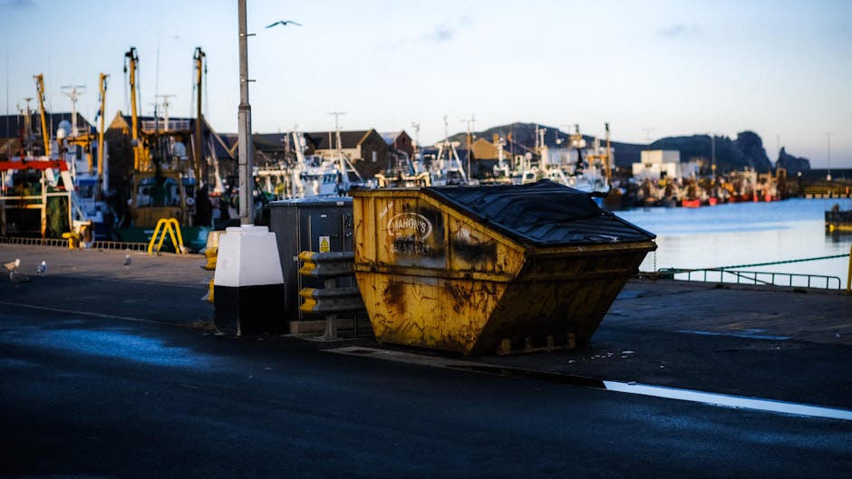 A large yellow industrial skip with visible rust and graffiti is situated on a paved dockside area next to a body of water. The skip is positioned near a pole and a metal utility box, with a protective white and black bollard nearby. Behind the skip, there is a wide view of a marina filled with various boats and yachts moored along the quay. The boats have masts, rigging, and equipment typical of fishing or leisure vessels. In the distance, there are low-rise buildings, some with pitched roofs, and a few lampposts along the waterfront. The scene is illuminated by natural daylight, suggesting either early morning or late afternoon, capturing the industrial and maritime environment typical of a port used for home relocation and furniture transport logistics. This setting reflects the process of loading or temporary storage, relevant to household removals services provided by Man with Van St Helier.