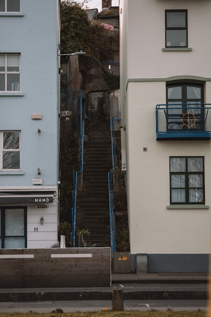 Image showing a steep outdoor staircase with wooden steps and blue metal railings, situated between two multi-storey residential buildings. The left building is light blue with multiple windows, while the right building is white with a small balcony featuring a black metal railing. The staircase leads up a hillside, partly obscured by bushes and trees, with some natural terrain visible at the top. The scene is captured during daylight hours, with shadows cast by the buildings. This setting illustrates a narrow access point often encountered in house removals and home relocation processes, where careful planning is needed for furniture transport and packing through constrained pathways. Occasionally, Man with Van St Helier would manage such challenging access routes during the loading and unloading phases of a move, ensuring careful handling of belongings along narrow lanes and staircases typical of the St Helier estate.