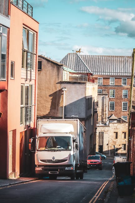 A man wearing a blue jacket and matching blue trousers is seen from behind, loading cardboard moving boxes into the rear of a white commercial van parked on a cobbled street in St Helier. The van's ba