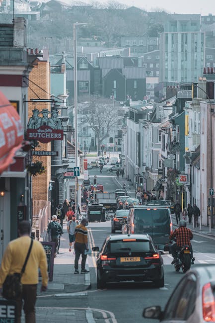 A busy urban street scene in St Helier showing a mix of pedestrians walking along the sidewalk, some carrying bags or backpacks, and others standing near shopfronts. Several vehicles, including a black car, a van, and a motorcycle, are moving or parked along the road. The street is lined with small retail shops, including a butchers shop with a sign reading 'BUTCHERS' and a sign for a 'FREEHOUSE.' Building facades feature various signs, shopfronts, and exterior lighting fixtures. The background includes residential and commercial buildings of different architectural styles with some taller structures visible further back, all under a cloudy daytime sky. This setting reflects the urban environment typical of house removals or furniture transport operations, which local companies like Man with Van St Helier might assist with during home relocation projects or packing and moving logistics in central St Helier.