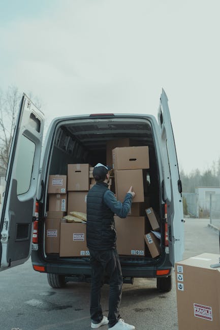 A man wearing a black jacket, grey trousers, and a black cap is standing at the open rear of a large white moving van, which is parked outdoors on a paved area. Inside the van, multiple brown cardboard boxes of varying sizes are stacked, some secured with packing tape and others resting directly on the van’s floor. The boxes are arranged to optimize space for home relocation purposes, with some positioned upright and others on their sides. The man is using his right hand to handle a medium-sized box, preparing to load or unload it from the vehicle. Several boxes are placed on the ground nearby, ready for transportation. The van is fitted with interior lining and features an open side door, indicating an ongoing loading or unloading process associated with packing, furniture transport, or moving logistics, with a background of an overcast sky and minimal outdoor structures from the vicinity of the premises where the move is taking place. Man with Van St Helier handles professional removals with careful packing and transport of household items.