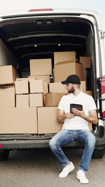 A man wearing a white t-shirt, blue jeans, white sneakers, and a black cap is seated on the edge of an open van's lowered rear door during a home relocation. Inside the van, numerous cardboard boxes of various sizes are stacked securely, some wrapped with protective plastic film and others with packing tape, prepared for furniture transport and moving logistics. The boxes are arranged in an organized manner, filling the space from the floor to near the ceiling, with a few larger boxes positioned at the bottom. The man is holding a tablet in both hands, possibly coordinating the packing or move, and the van is parked on a paved surface. The setting suggests a loading process associated with professional removals and moving services provided by Man with Van St Helier, supporting efficient packing and moving operations during house removals in the local area.