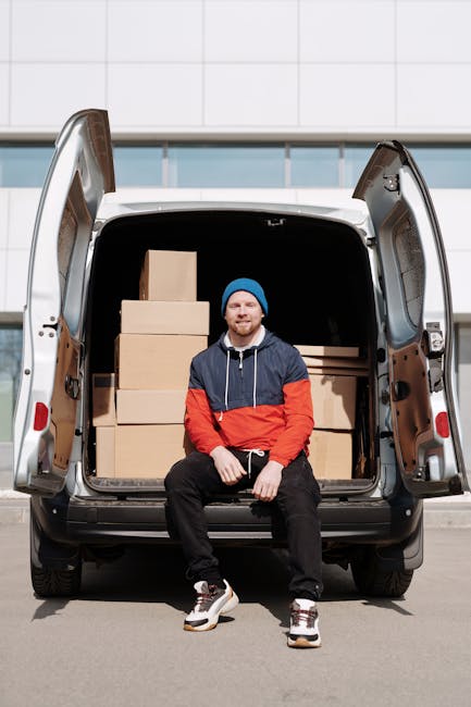 A man wearing a black jacket, grey trousers, and a black cap is standing at the open rear of a large white moving van, which is parked outdoors on a paved area. Inside the van, multiple brown cardboard boxes of varying sizes are stacked, some secured with packing tape and others resting directly on the van’s floor. The boxes are arranged to optimize space for home relocation purposes, with some positioned upright and others on their sides. The man is using his right hand to handle a medium-sized box, preparing to load or unload it from the vehicle. Several boxes are placed on the ground nearby, ready for transportation. The van is fitted with interior lining and features an open side door, indicating an ongoing loading or unloading process associated with packing, furniture transport, or moving logistics, with a background of an overcast sky and minimal outdoor structures from the vicinity of the premises where the move is taking place. Man with Van St Helier handles professional removals with careful packing and transport of household items.