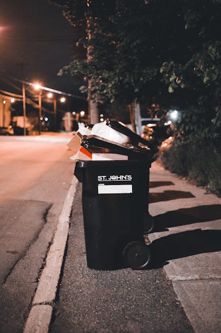 A black wheelie bin labeled 'St. John's' positioned on a dimly lit street at night, with its lid open revealing cardboard boxes, paper, and plastic packaging materials inside. The bin is located on the pavement near the curb, with shadows cast by nearby trees and streetlights. In the background, there are traffic lights, streetlights, and foliage, indicating an urban residential area. This image exemplifies waste disposal during the home relocation process, which is a key aspect of moving logistics. The presence of packaging waste underscores the importance of proper disposal laws, such as those referenced in the guide on waste disposal laws for St Helier moves by [COMPANY_NAME], a professional removals service specializing in house relocations, furniture transport, and packing and moving services in the area.