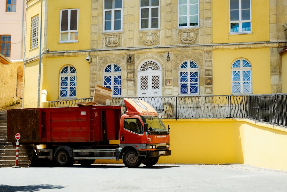 A red waste collection truck is parked on a paved street in front of a yellow residential building with decorative stonework around the arched windows and a white ornate front door. The truck is positioned near a yellow retaining wall with a black metal railing on top, and a cardboard box is resting on top of the truck's cargo area. The truck appears to be in the process of loading or unloading household waste or building materials, with part of the cargo area open and accessible. The environment is well-lit with natural daylight, and adjacent there are exterior stairs leading down the side of the building. This scene illustrates the waste disposal process associated with home relocation or furniture transport, which is relevant to house removals and moving services by Man with Van St Helier.